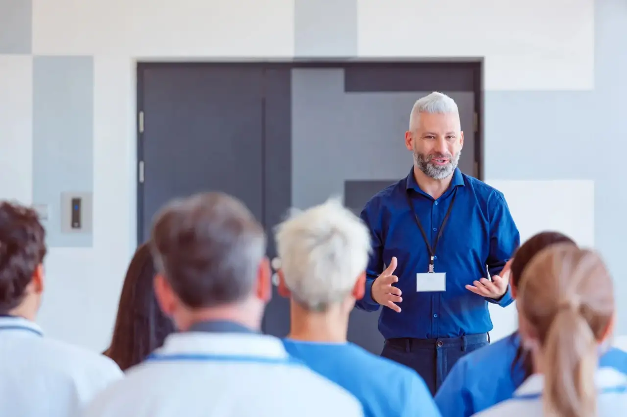 Male instructor in blue shirt with lanyard speaking to a seated older-adult audience.