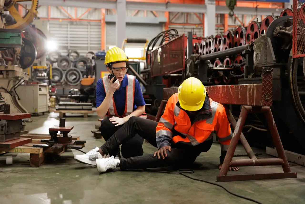 Factory workers, one in orange hi-vis jacket and yellow hard hat sitting with a colleague.