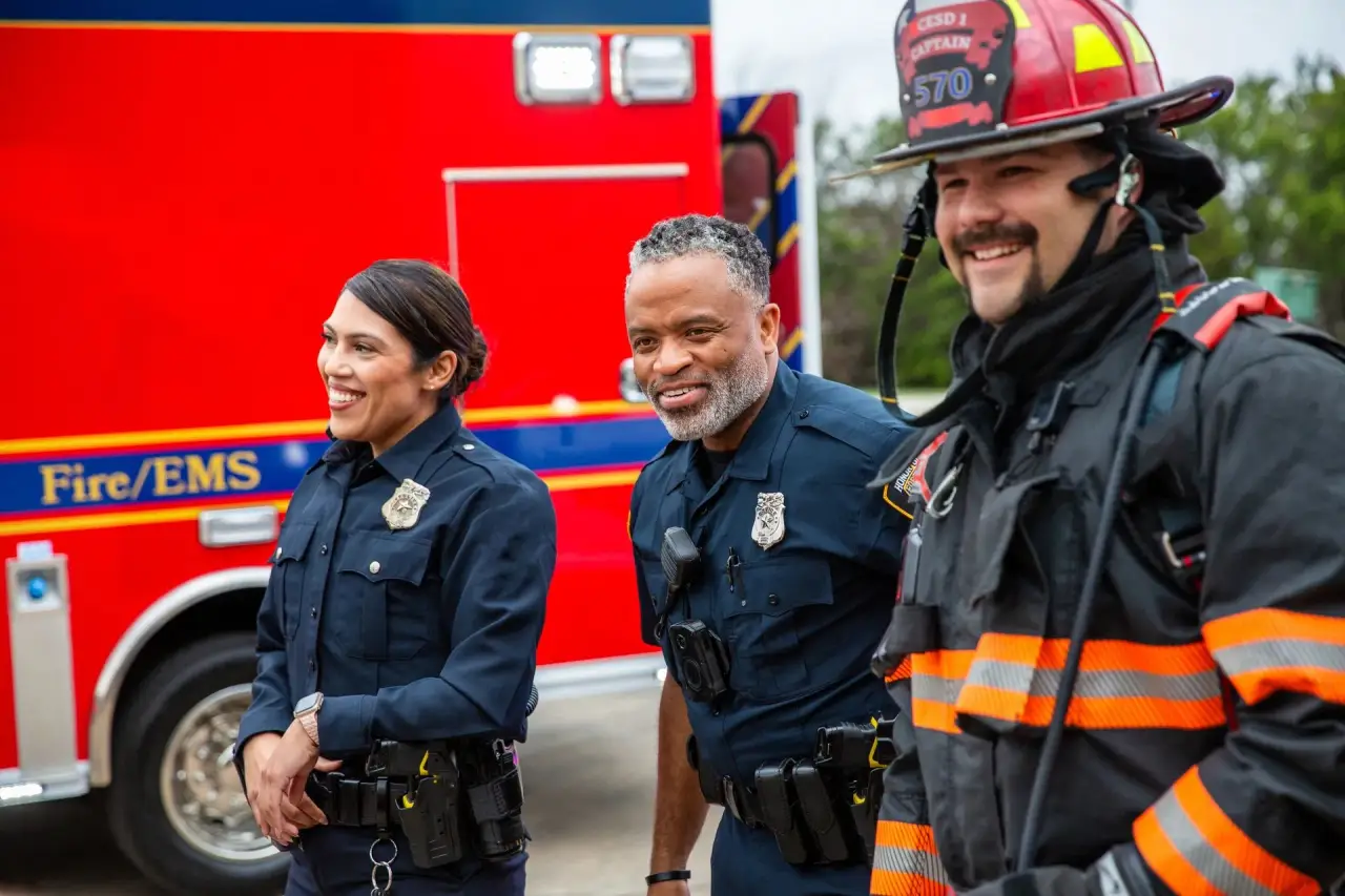 Two uniformed first responders (police/EMS, dark blue uniforms) smiling, red emergency vehicle behind.