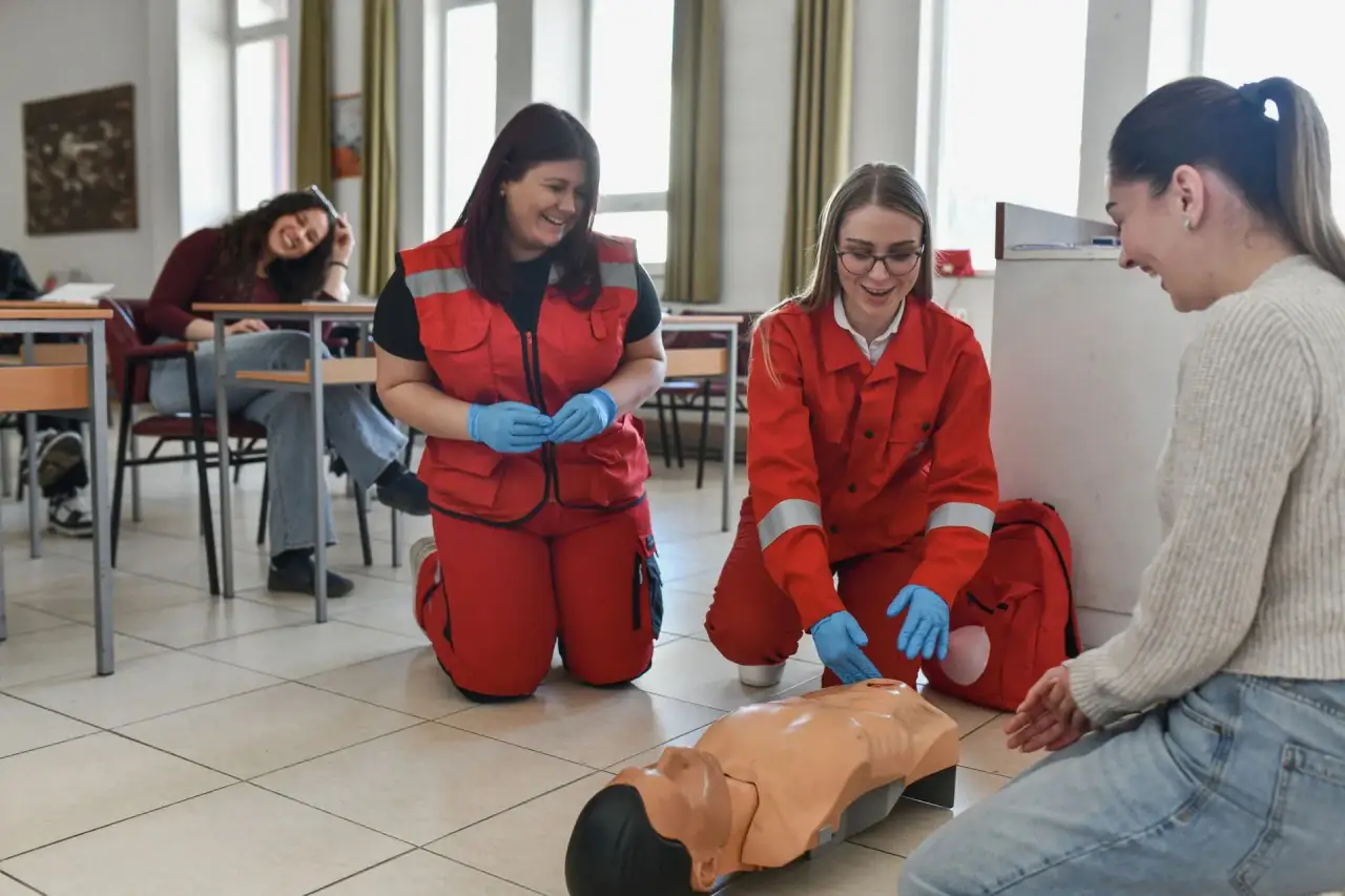 Responders in red uniforms practicing CPR on a manikin in a classroom while others watch.